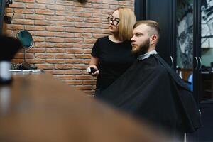 Stylish man sitting barber shop Hairstylist Hairdresser Woman cutting his hair Portrait handsome happy young bearded caucasian guy getting trendy haircut Attractive barber girl working serving client. photo