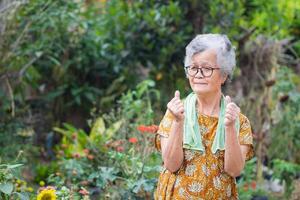 Portrait of an elderly woman exercises by gesturing raise both arms, smiling, and looking at the camera while standing in a garden. Space for text. Concept of aged people and healthcare photo