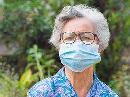 un mayor mujer vistiendo un quirúrgico máscara y mirando a el cámara mientras en pie en un jardín. máscara para proteger coronavirus, COVID-19, y más. concepto de Envejecido personas y cuidado de la salud foto