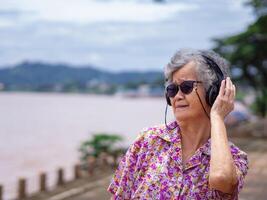 Portrait of a happy elderly woman wearing wireless headphones listening to music while standing side the river. Space for text. Concept of aged people and relaxation photo