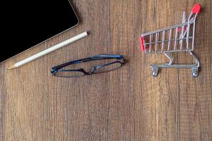 Top view of a pencil, glasses, mini shopping cart, and tablet on wooden table in the office. Space for text. Wood texture background. Business and e-commerce concept photo