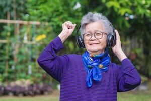 mayor mujer con corto gris pelo vistiendo lentes y inalámbrico auriculares escuchando a un canción, sonriente mientras en pie en un jardín. concepto de Envejecido personas y relajación foto