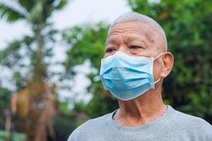 retrato de un mayor hombre vistiendo un cara máscara y mirando lejos mientras en pie en un jardín. máscara para proteccion virus, COVID-19, coronavirus, bacterias concepto de Envejecido personas y cuidado de la salud foto