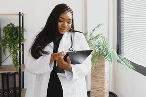 retrato de hermosa sonriente hembra africano americano médico en pie en médico oficina. salud cuidado concepto, médico seguro, Copiar espacio foto