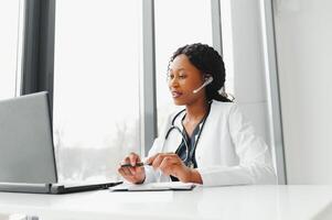 African American woman doctor working at her office online using portable inormation device. Telemedicine services. Primary care consultations, psychotherapy, emergency services photo