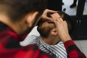 grave barbado hombre consiguiendo barba Corte de pelo con un Derecho maquinilla de afeitar por Barbero mientras sentado en silla a peluquería. barbería tema. foto