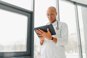 Mature african doctor using digital tablet in corridor . Portrait of confident male doctor using tablet computer in clinic with copy space. Successful smiling doctor in labcoat wearing stethoscope photo