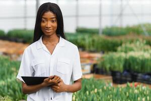 innovación tecnología para inteligente granja sistema. sonriente africano americano niña cheques flores en invernadero con tableta en su manos, Dom llamarada foto