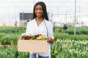 Spring and seedlings. Smiling african american girl in apron carries a box with young plants on flowers background in interior of greenhouse photo