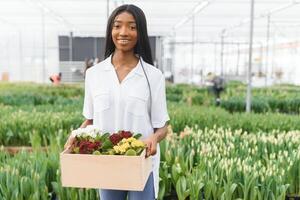 Spring and seedlings. Smiling african american girl in apron carries a box with young plants on flowers background in interior of greenhouse photo