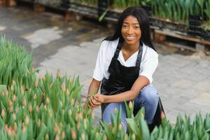 Beautiful young smiling african american girl, worker with flowers in greenhouse. Concept work in the greenhouse, flowers. photo