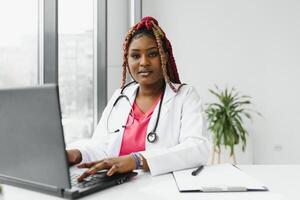 medicine, online service and healthcare concept - happy smiling african american female doctor or nurse with headset and laptop having conference or call at hospital. photo