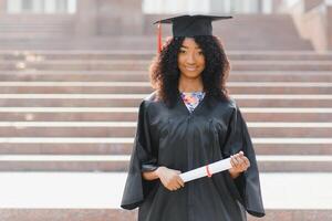 alegre afro americano hembra graduado en pie en frente de Universidad edificio foto