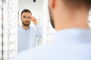 Choosing Glasses Concept. Portrait of handsome bearded guy picking new specs at optical shop, looking at mirror photo