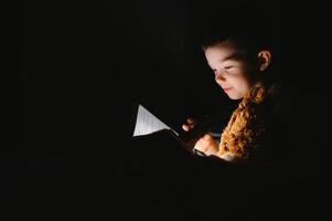 Boy with flashlight reading book under blanket at home. photo