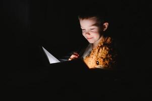 Portrait of cute little boy reading in bed with flashlight in dark room, enjoying fairytales. photo