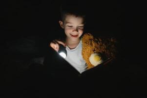 niño leyendo libro en cama. niños leer a noche. pequeño chico con hada cuento libros en dormitorio . educación para joven niños. hora de acostarse historia en el noche. linda niño debajo cobija en oscuro habitación con lámpara foto