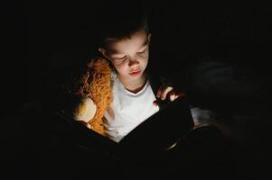 chico con Linterna leyendo libro debajo cobija a hogar. foto