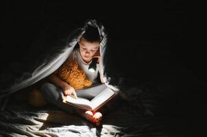 Boy with flashlight reading book under blanket at home. photo