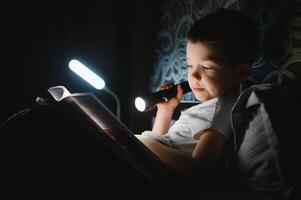 Boy with flashlight reading book under blanket at home. photo