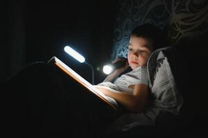 Portrait of cute little boy reading in bed with flashlight in dark room, enjoying fairytales. photo