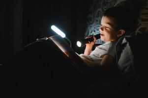 Reading book and using flashlight. Young boy in casual clothes lying down near tent at evening time photo