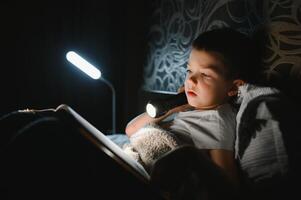 niño leyendo libro en cama. niños leer a noche. pequeño chico con hada cuento libros en dormitorio . educación para joven niños. hora de acostarse historia en el noche. linda niño debajo cobija en oscuro habitación con lámpara foto