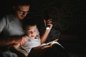 padre y hijo con Linterna leyendo libro debajo cobija a hogar. foto