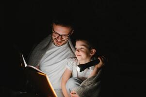 Happy family reading bedtime story under blanket in evening. Father and son spend time together. Father's Day photo