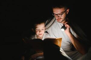 Happy family reading bedtime story under blanket in evening. Father and son spend time together. Father's Day photo