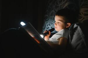 Portrait of cute little boy reading in bed with flashlight in dark room, enjoying fairytales. photo