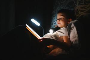 Reading book and using flashlight. Young boy in casual clothes lying down near tent at evening time. photo