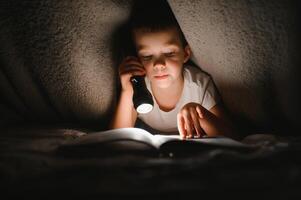 niño leyendo libro en cama. niños leer a noche. pequeño chico con hada cuento libros en dormitorio . educación para joven niños. hora de acostarse historia en el noche. linda niño debajo cobija en oscuro habitación con lámpara foto