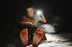 niño leyendo libro en cama. niños leer a noche. pequeño chico con hada cuento libros en dormitorio . educación para joven niños. hora de acostarse historia en el noche. linda niño debajo cobija en oscuro habitación con lámpara foto