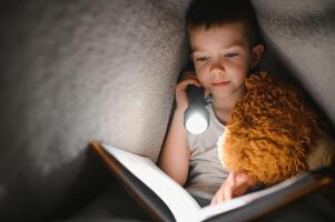 un chico de 5-6 años antiguo es leyendo un libro en el noche en el oscuro debajo un cobija con un juguete oso foto