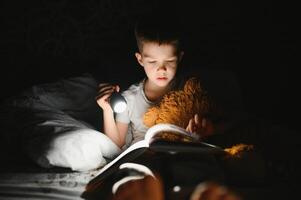 Portrait of cute little boy reading in bed with flashlight in dark room, enjoying fairytales. photo