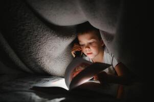 Boy with flashlight reading book under blanket at home photo
