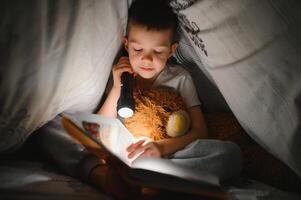 un chico de 5-6 años antiguo es leyendo un libro en el noche en el oscuro debajo un cobija con un juguete oso. foto