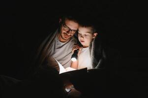 Happy family reading bedtime story under blanket in evening. Father and son spend time together. Father's Day photo