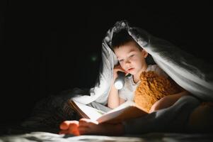 chico con Linterna leyendo libro debajo cobija a hogar. foto