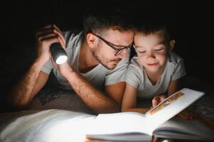 Happy family reading bedtime story under blanket in evening. Father and son spend time together. Father's Day photo