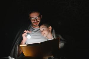 padre y hijo con Linterna leyendo libro debajo cobija a hogar. foto