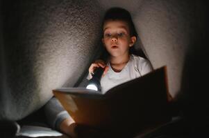 niño leyendo libro en cama. niños leer a noche. pequeño chico con hada cuento libros en dormitorio . educación para joven niños. hora de acostarse historia en el noche. linda niño debajo cobija en oscuro habitación con lámpara foto