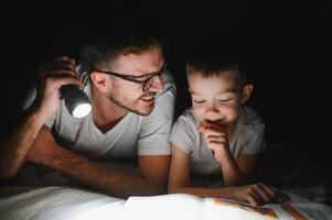 Happy family reading bedtime story under blanket in evening. Father and son spend time together. Father's Day photo