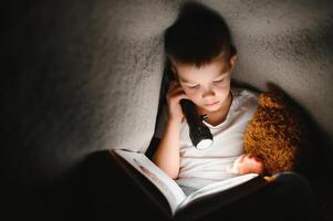 Portrait of cute little boy reading in bed with flashlight in dark room, enjoying fairytales. photo