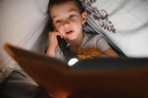 Reading book and using flashlight. Young boy in casual clothes lying down near tent at evening time photo