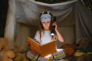 niño niña leyendo con libro y Linterna y osito de peluche oso en carpa. antes de yendo a cama foto