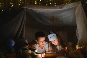 Two children with flashlight read a book under a blanket as a tent photo