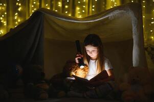 child girl reading with book and flashlight and teddy bear in tent. before going to bed photo