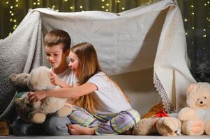 Two little child play at home in the evening to build a camping tent to read books with a flashlight and sleep inside. photo